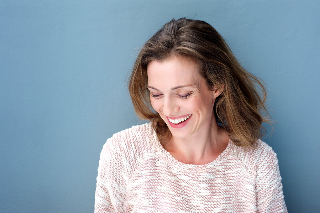woman in pink sweater laughs, standing against a blue backdrop