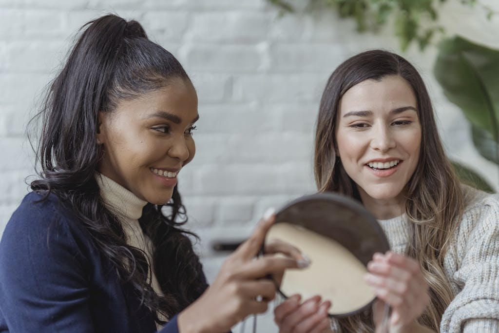 Happy young ethnic woman giving mirror to female client after applying makeup